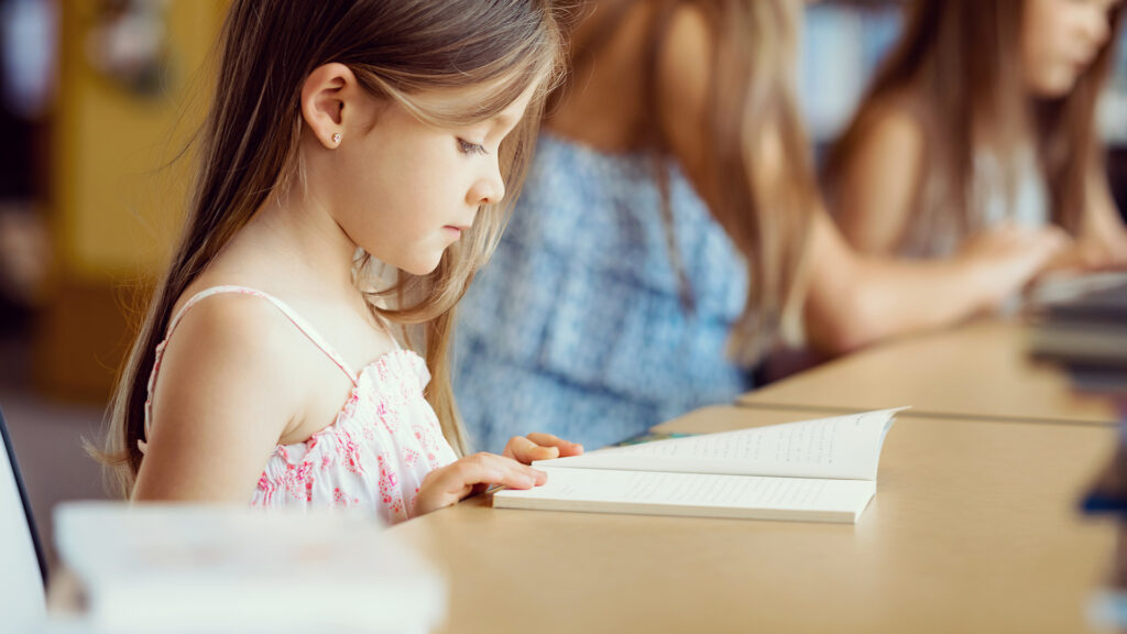 Elementary school student is sitting at a table in a library reading a book