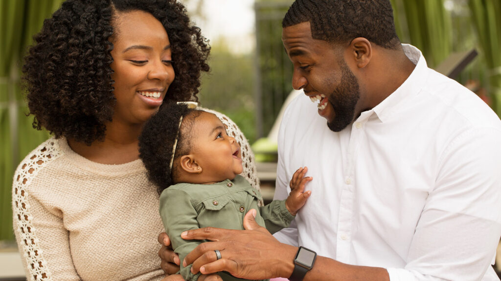 A mother and father hold a baby and engage the baby in talking and playing with sounds.
